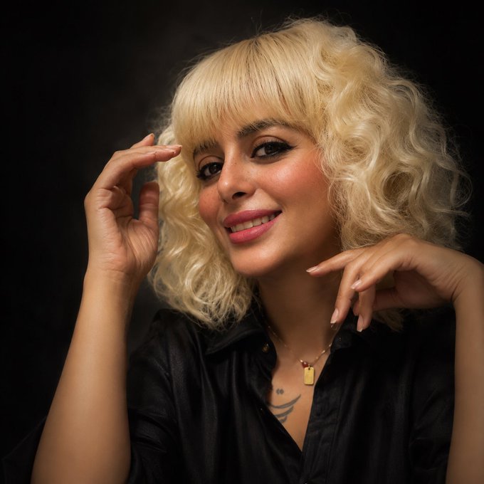 A studio portrait shows a smiling woman with curly blonde hair, dark eyeliner, and a black satin shirt, posing with her hands near her face against a black background.