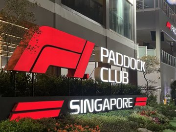 The image shows the official F1 Paddock Club signage in Singapore, featuring the iconic red F1 logo alongside the words "PADDOCK CLUB" and "SINGAPORE" displayed prominently against a building exterior. This represents the exclusive hospitality venue where the author and their friends watched the Singapore Grand Prix, despite not attending in person.