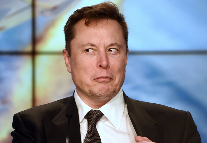 A close-up photograph of Elon Musk wearing a dark suit, white shirt, and patterned tie, looking sideways with a serious expression, photographed at what appears to be a formal event or press conference.