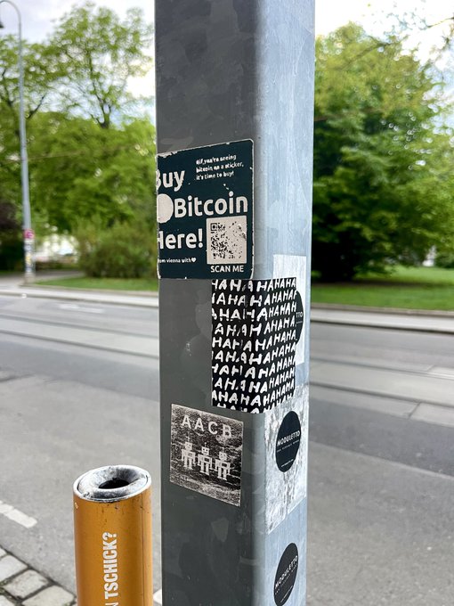 A street pole covered in various stickers, including a Bitcoin sticker with a QR code, a sticker with 'AHA' repeated across it, and an 'AACB' sticker, with a yellow bollard and trees visible in the background.