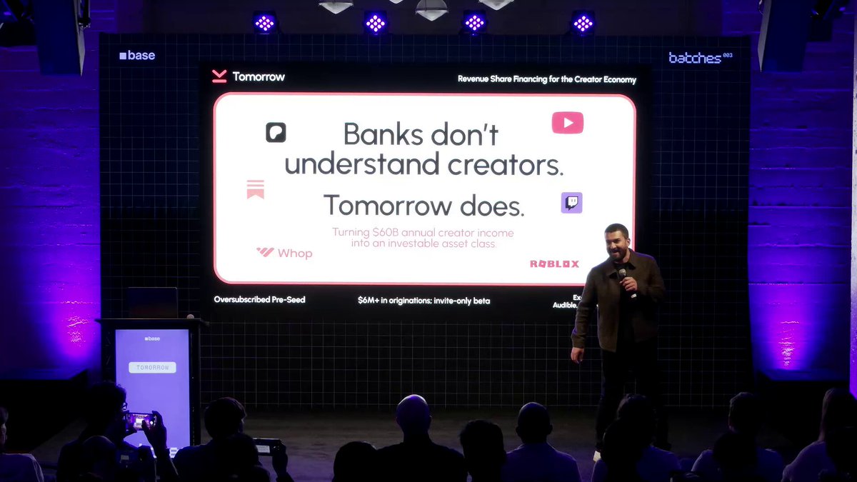 The image shows a man standing behind a podium at an event, giving a speech or presentation. The audience consists of several people sitting in front of him, attentively listening to his words. In the background, there are purple lights illuminating the stage, creating a focused atmosphere for the event. On the screen behind the speaker, there is a large white sign with black text that reads "→ @tomorrowloans Stablecoin lending to creators, secured against verifiable Google AdSense using zkTLS". The image also contains some text in the bottom right corner which seems unrelated to the main scene but might be related to the event or speaker.