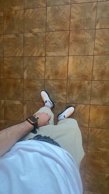 The image shows a person wearing white sneakers, khaki pants, and a white t-shirt standing on a brown floor with their feet crossed over each other. The person has a watch on their left wrist and appears to be looking down at something in front of them. There is also an Apple logo visible on the person's wristwatch.