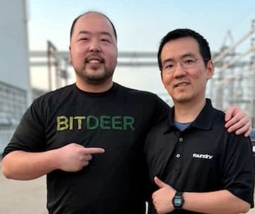 Two men stand together, one wearing a black 'BITDEER' t-shirt and the other in a 'Rudy' polo shirt, pointing at the logo.