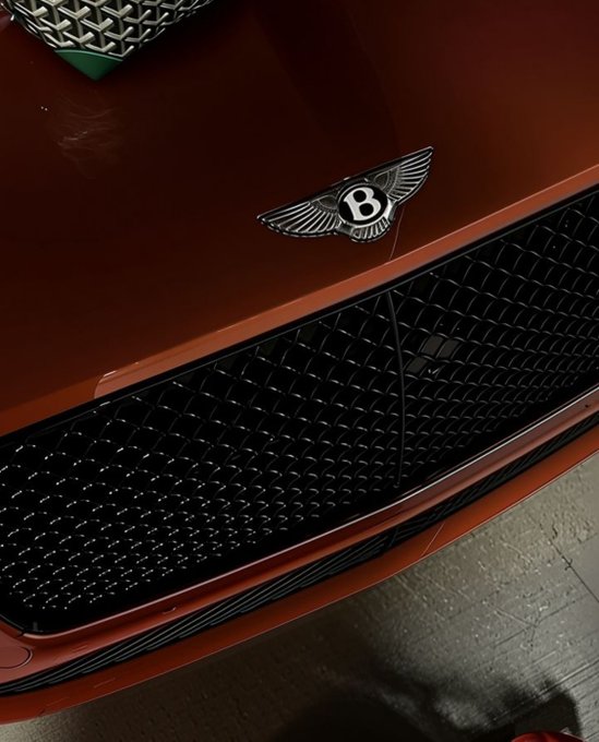 A close-up photo of a red Bentley vehicle's front grille featuring the iconic winged 'B' emblem on the hood above the signature mesh grille.