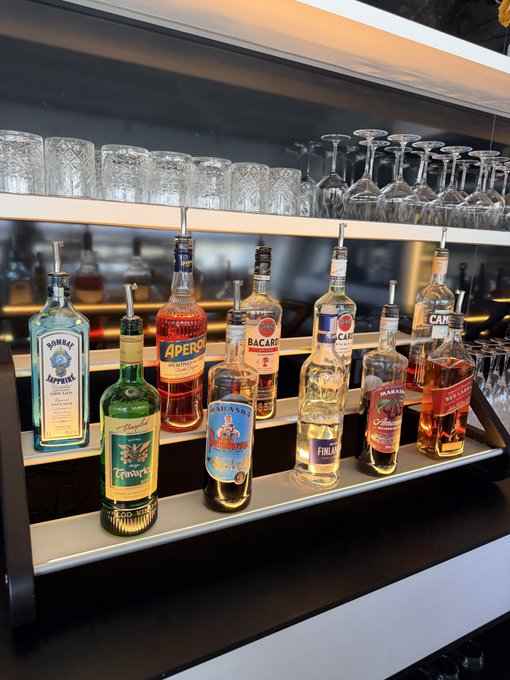 A bar setup with a row of liquor bottles including Bombay Sapphire, Aperol, Bacardi, Tanqueray, and others, equipped with pour spouts, displayed on a shelf with glassware visible on a rack above.