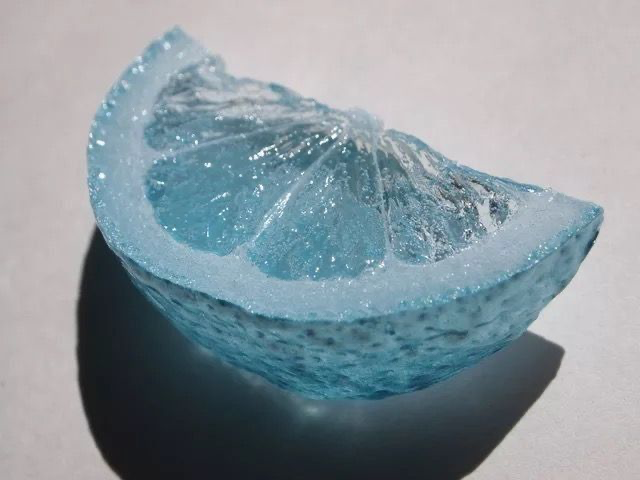 A close-up of a translucent, light blue resin object shaped and textured like a citrus slice, casting a shadow on a white surface.