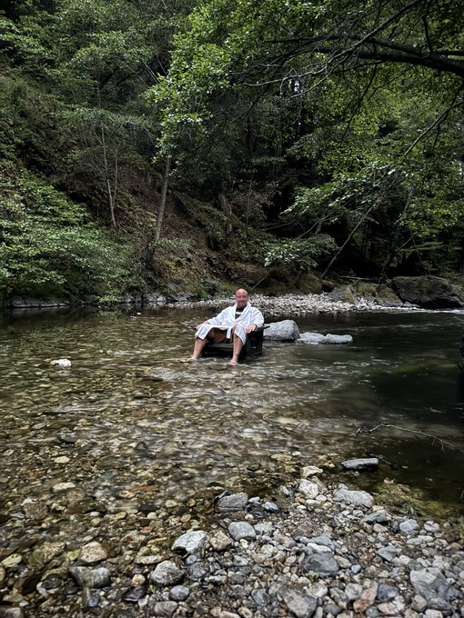 The image shows a man sitting in a chair at the edge of a river, wearing a robe or bathrobe. The water appears to be calm, with rocks visible around him. The caption suggests that this scene might be related to a lifestyle or travel blog post about living on a river and enjoying simple pleasures like taking baths or showers outdoors.