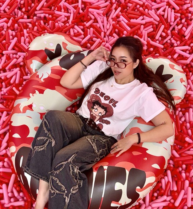 A woman wearing a white 'SEOUL' Betakkuma t-shirt and ripped dark jeans relaxes in a camouflage-patterned bean bag surrounded by red and pink foam noodles, wearing red-tinted glasses.