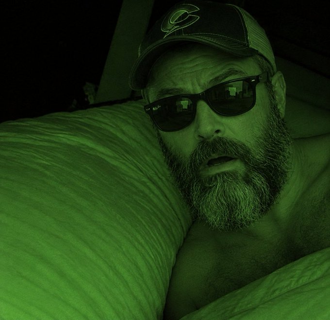 A nighttime photo with green night vision showing a bearded man wearing a baseball cap with a 'C' logo and sunglasses, lying in bed.