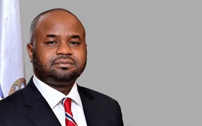A man in a black suit with a red, white, and blue striped tie poses for an official portrait against a gray background, with a partially visible flag behind him—likely representing a government or institutional figure, such as an official from the Nigeria Revenue Service (NRS), in connection with the post about NRS guidelines on virtual assets taxation.