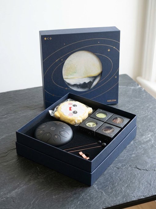 A dark blue gift box from "MSX.COM" featuring an open lid decorated with a moon and orbital ring design sits on a black surface, displaying its contents: a round black percussion instrument (tongue drum), a smiley-faced cookie, an assortment of moon-themed chocolates, and a small mallet.