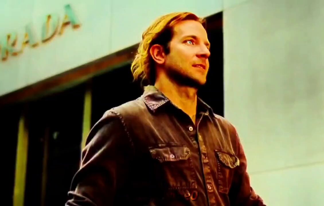 The image features a man wearing a brown jacket with his hair styled long. He appears to be looking off to the side or walking away from the camera, possibly engaged in an activity outside of the frame. The background includes a building and a sign that reads "RAD".