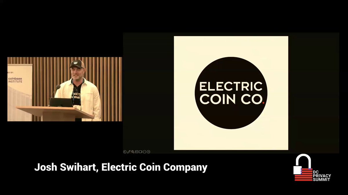 The image features a man standing behind a podium at an event, giving a presentation about electric coin company Zcash. The background of the image shows a black screen displaying the logo for Electric Coin Company along with some text. The main focus of the image is on the speaker and his presentation.
