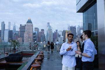 A group of professionals gathered at a rooftop event, engaged in conversation with the illuminated Manhattan skyline as a backdrop. The image captures the social atmosphere of the DeFi and finance networking dinner mentioned in the post, though no specific text or logos are visible in the scene itself.