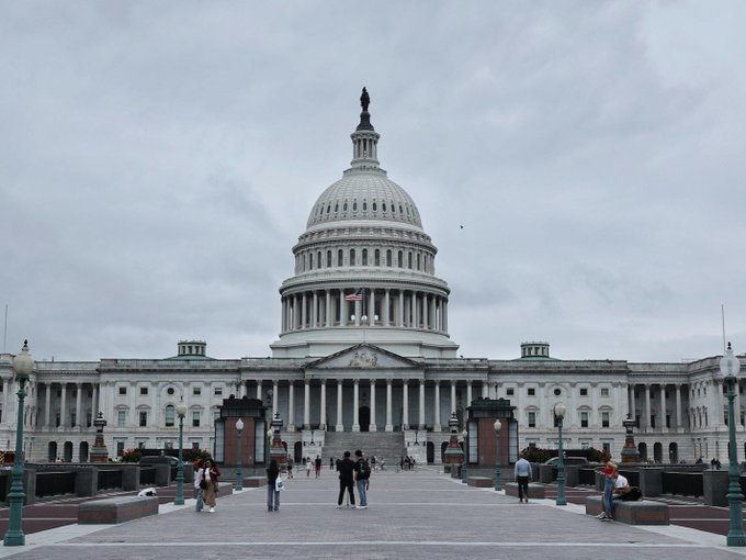 The image shows the United States Capitol Building in Washington, D.C., with visitors walking around the grounds on an overcast day, with an American flag visible on the dome. The image relates to the post as it depicts the seat of the U.S. Senate, where Senator Elizabeth Warren made her comments opposing the CLARITY Act and advocating for alternative crypto legislation.