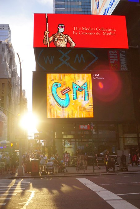 The image shows a busy city street with people walking around, cars driving by, and tall buildings lining the street. The sun is shining brightly, creating shadows that add depth to the scene. In the foreground of the photo, there are two large billboards - one red and one yellow. One billboard features an illustration of a man holding a sword, while the other has a cartoon character on it. A person can be seen walking near these billboards, possibly admiring them or going about their day.