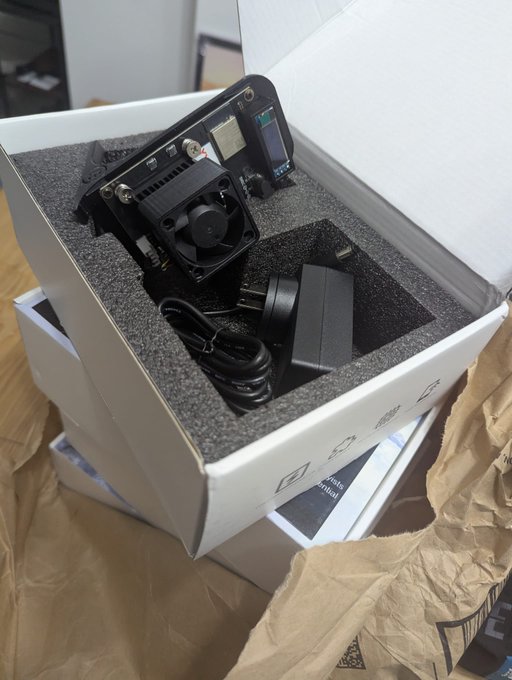 An electronic device with a cooling fan and power supply unit packaged in foam inside a cardboard box, sitting on brown packing paper.