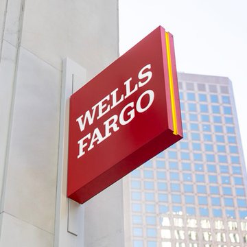 A red Wells Fargo sign is mounted on the exterior of a modern building, with a glass office tower visible in the background. The image visually represents the bank at the center of the news about its potential crypto trading liquidity partnership with Payward, the parent company of Kraken.