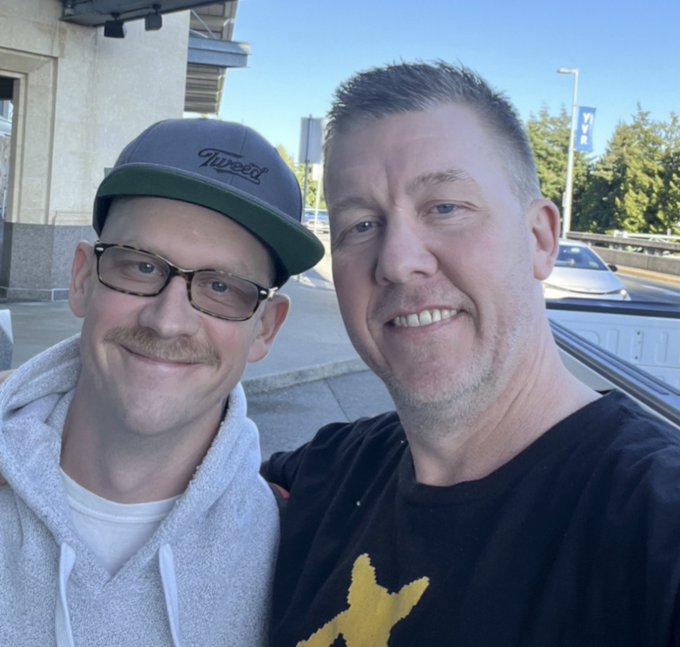 Two men pose for a selfie outdoors, one wearing a gray "Flyers" cap and glasses, the other in a black t-shirt with a yellow design.