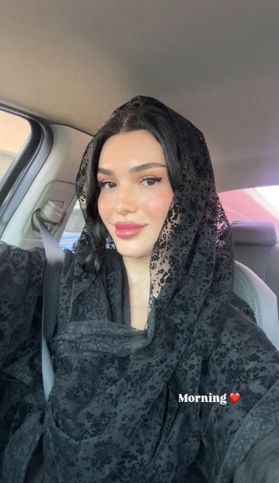 A selfie of a woman wearing a black lace hijab and full makeup, sitting in a car with a seatbelt on, with the text 'Morning' and a heart emoji overlaid on the image.