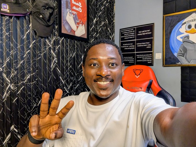 A man smiles warmly while giving a peace sign in his room, which is decorated with framed artwork and a colorful gaming chair. The cheerful selfie pairs perfectly with his "Good Morning Folks" post, capturing a grateful and blessed Sunday vibe.