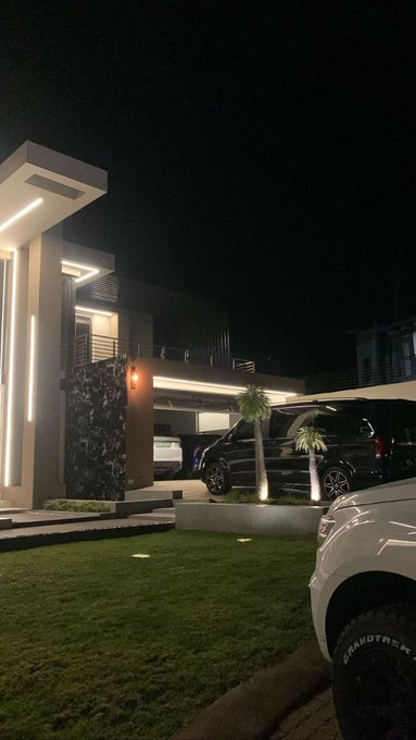 A nighttime exterior view shows a modern luxury home with illuminated architecture, a covered garage housing vehicles, and palm trees in the landscaping.
