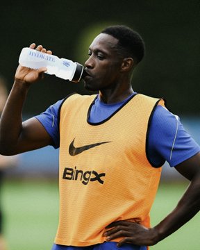 A soccer player in a blue shirt and orange Nike training bib with 'BINX' printed on it drinks from a clear water bottle labeled 'maeve.' on a training pitch.