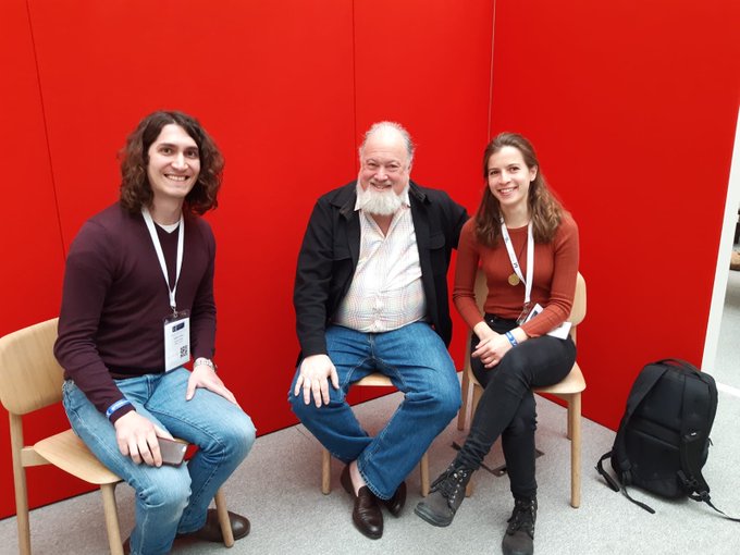 Three people—a young man, an older bearded man, and a young woman—sit smiling on wooden chairs against a bright red wall, all wearing lanyards.