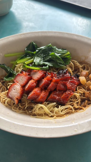 A bowl of egg noodles topped with sliced Chinese barbecue pork (char siu) and blanched leafy green vegetables, served on a teal table.