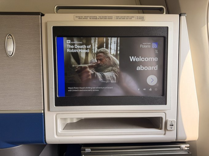 An airplane seatback entertainment screen displays the United Airlines welcome message along with a promotion for 'The Death of Robin Hood' on United's Polaris 1L.
