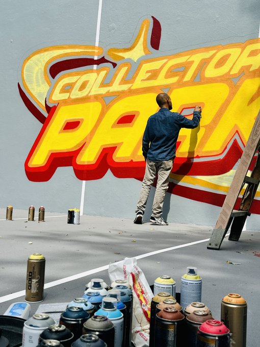 An artist in a denim jacket spray-paints colorful graffiti art reading 'COLLECTOR PARK' on a large gray wall, with numerous spray paint cans scattered in the foreground.