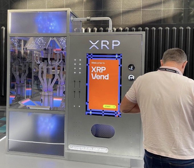A man interacts with an XRP-branded vending machine labeled "Welcome to XRP Vend" with a "START" button, featuring a transparent display showcasing the mechanism and illuminated by blue lighting. The image directly illustrates the post's excitement about an XRP-compatible ATM being demonstrated at the Permissionless conference, where users can insert cash to receive XRP tokens.