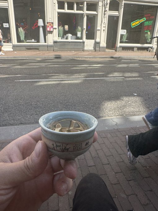A first-person view shows a hand holding a small ceramic cup of sake or rice wine while walking past storefronts on a brick-paved sidewalk.