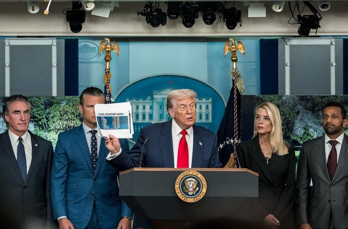 President Donald Trump speaks at a White House podium holding up a chart titled 'TOTAL WORKER A K-8', flanked by other officials including FBI Director Kash Patel and Attorney General Pam Bondi.