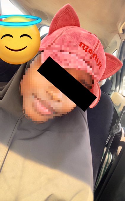 A selfie of a person wearing a pink Reebok cap with cat ears, their face partially obscured by a black censor bar and an angel emoji overlay, taken inside a car.