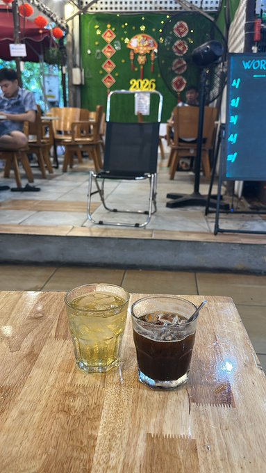 Two glasses of iced beverages on a wooden table at a café, with a decorative green backdrop featuring '2026' and Chinese characters visible in the background.