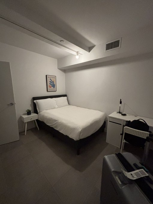 A dimly lit, minimalist bedroom featuring a queen-sized bed with white linens, a small nightstand, and a desk—likely in a co-living space or shared apartment. In the foreground, a suitcase with a luggage tag sits on the floor, suggesting the occupant has just arrived, fitting the post's theme of meeting new roommates in a new living arrangement.