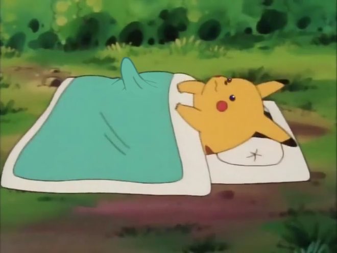 Pikachu lying on the ground wrapped in a teal blanket on a white mat in a grassy outdoor setting.