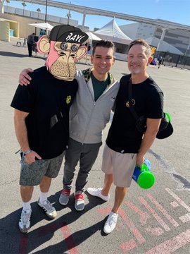 Three men pose together outdoors, with one wearing a cartoonish oversized mask resembling a 'PAW Patrol' character, while the other two smile beside him.