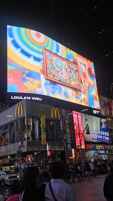 A bustling nighttime city scene dominated by a massive illuminated digital billboard labeled 'LOULAN WEU,' with a McDonald's sign and T-Mobile 'Gifts' advertisements visible below.