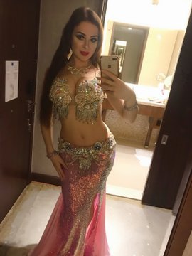 A woman poses for a mirror selfie in an ornately embellished, jewel-toned belly dance costume, capturing the glamorous "before" lifestyle of performing in luxury hotels abroad before transitioning to remote work in crypto. The luxurious indoor setting reflects the five-star hotel life described in her post.