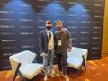 Two men pose together for a photo on an orange carpet at what appears to be a conference or event venue.
