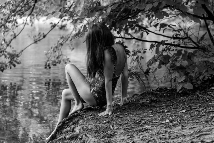 The image features a woman sitting on a rock by a body of water, possibly a lake or pond. She has her back turned towards the camera, giving us a view of her from behind. The photo appears to be black and white, which adds an artistic touch to the scene. There is no text present in the image, but there are several words written on it that read "Touching grass with 
Mermaid before sleep Thank you, my dear @thePrettyProof for adding it to your soulful collection!".