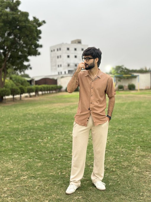 A young man wearing sunglasses, a tan button-up shirt, and cream-colored pants stands on a grassy field, posing thoughtfully with his hand near his face.