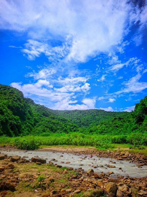 A scenic landscape featuring a rocky stream in the foreground, lush green hills in the midground, and a bright blue sky with scattered white clouds.