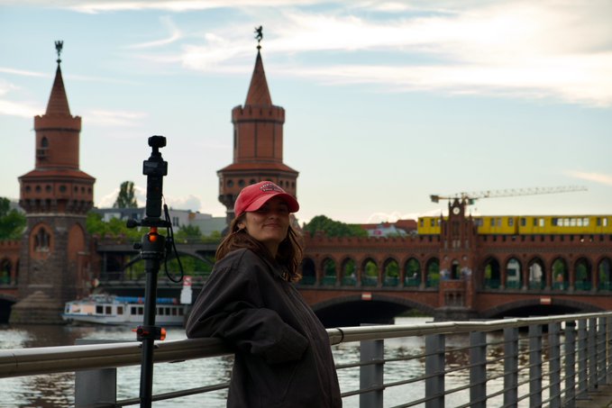 The image features a woman standing near a metal railing, wearing a red hat and a black jacket. She appears to be posing for a photo or video shoot. In the background, there are two towers with spires that can be seen against the sky. The cityscape suggests that they might have traveled from another country to attend an event in Berlin.
