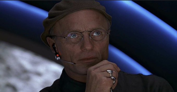 A contemplative man wearing round wire-rimmed glasses, a beige cap, and a headset with a glowing red light rests his chin on his hand, which bears a silver skull ring, against a backdrop of blue diagonal panels. The image appears to humorously suggest the thoughtful, almost calculating demeanor needed for apes (Wall Street traders) to settle down with some steady, organized "bookshelf tin" rather than chaotic market action.