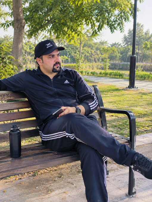 A man in a dark Adidas tracksuit and Reebok cap sits relaxed on a wooden park bench, holding a phone next to a black water bottle.
