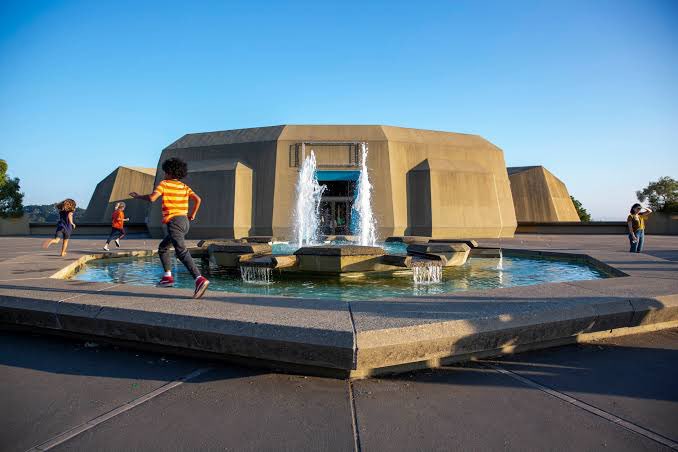 Children run around a tiered fountain in front of a brutalist-style concrete building under a clear blue sky.