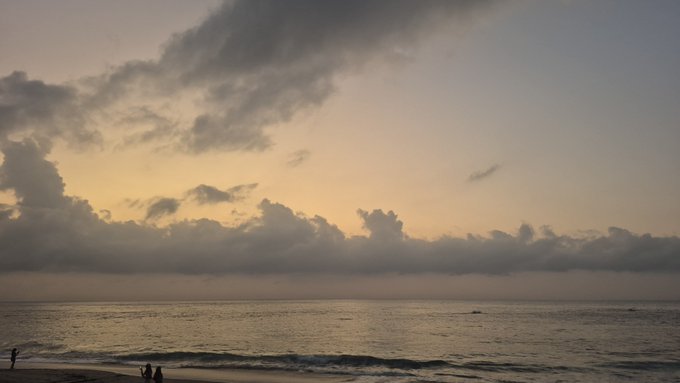 A serene ocean view at sunset or sunrise, featuring silhouetted clouds and a few small figures standing along the shoreline.